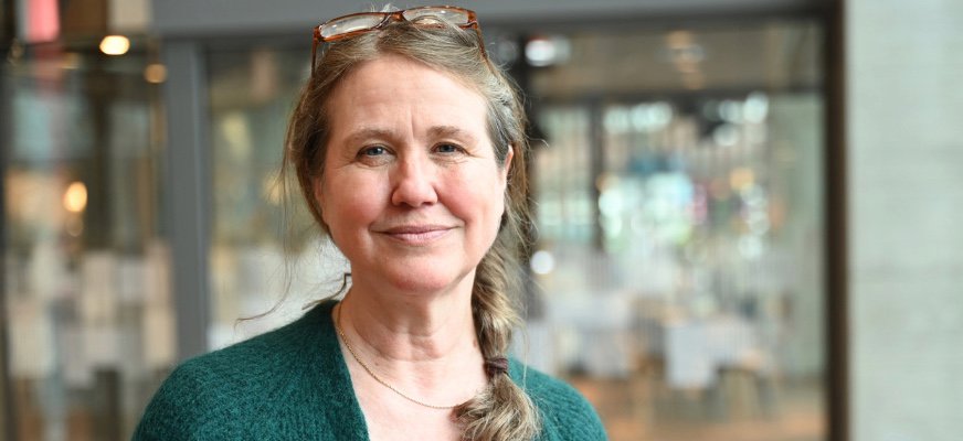 Lotta Gränse har långt hår i en fläta på ena sidan och glasögon placerade på huvudet. Hon bär en grön kofta, tittar in i kameran och ler.