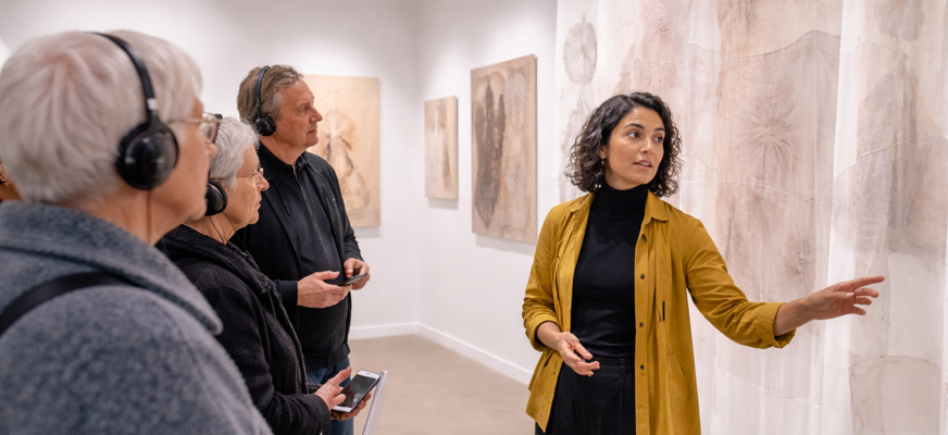 En guide visar en utställning för 3 personer som har hörlurar för syntolkning