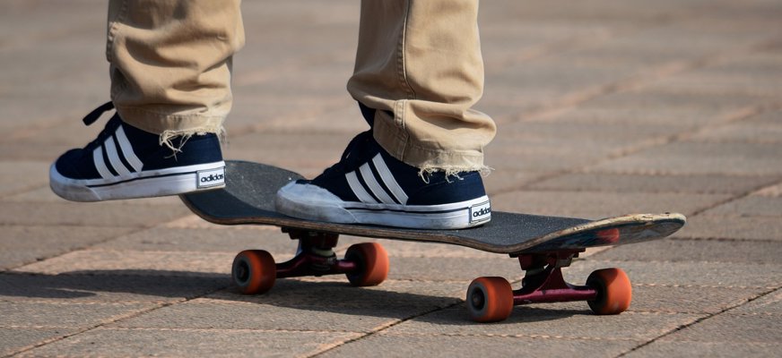 En inzoomad bild på en skateboard, ett par adidasskor och ljusa byxben.