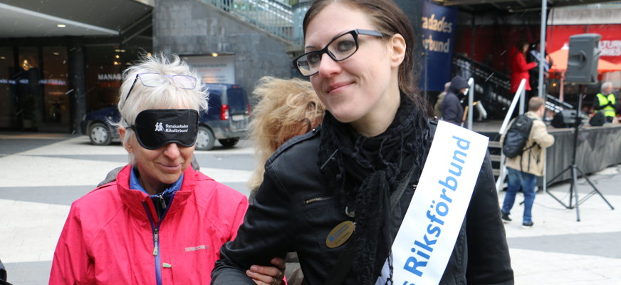 Leende kvinna i svartfärgat hår, glasögon och ordensband med SRF:s logotyp ledsagar äldre kvinna med ögonbindel på org.