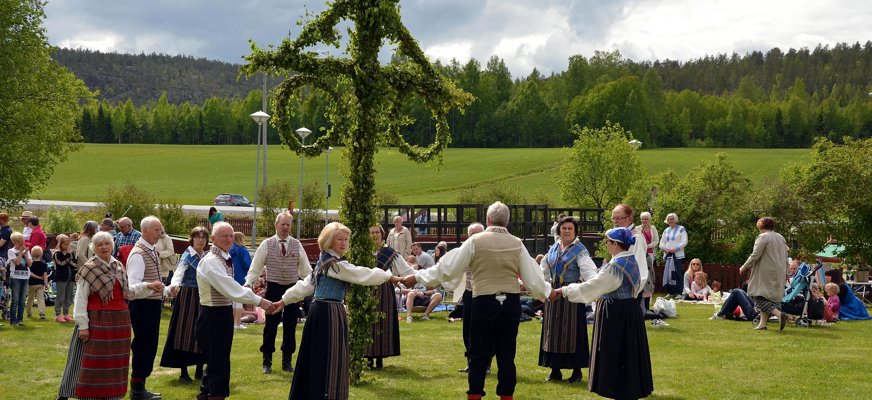 Bilden visar folkdräktsklädda personer som dansar i en ring runt en midsommarstång