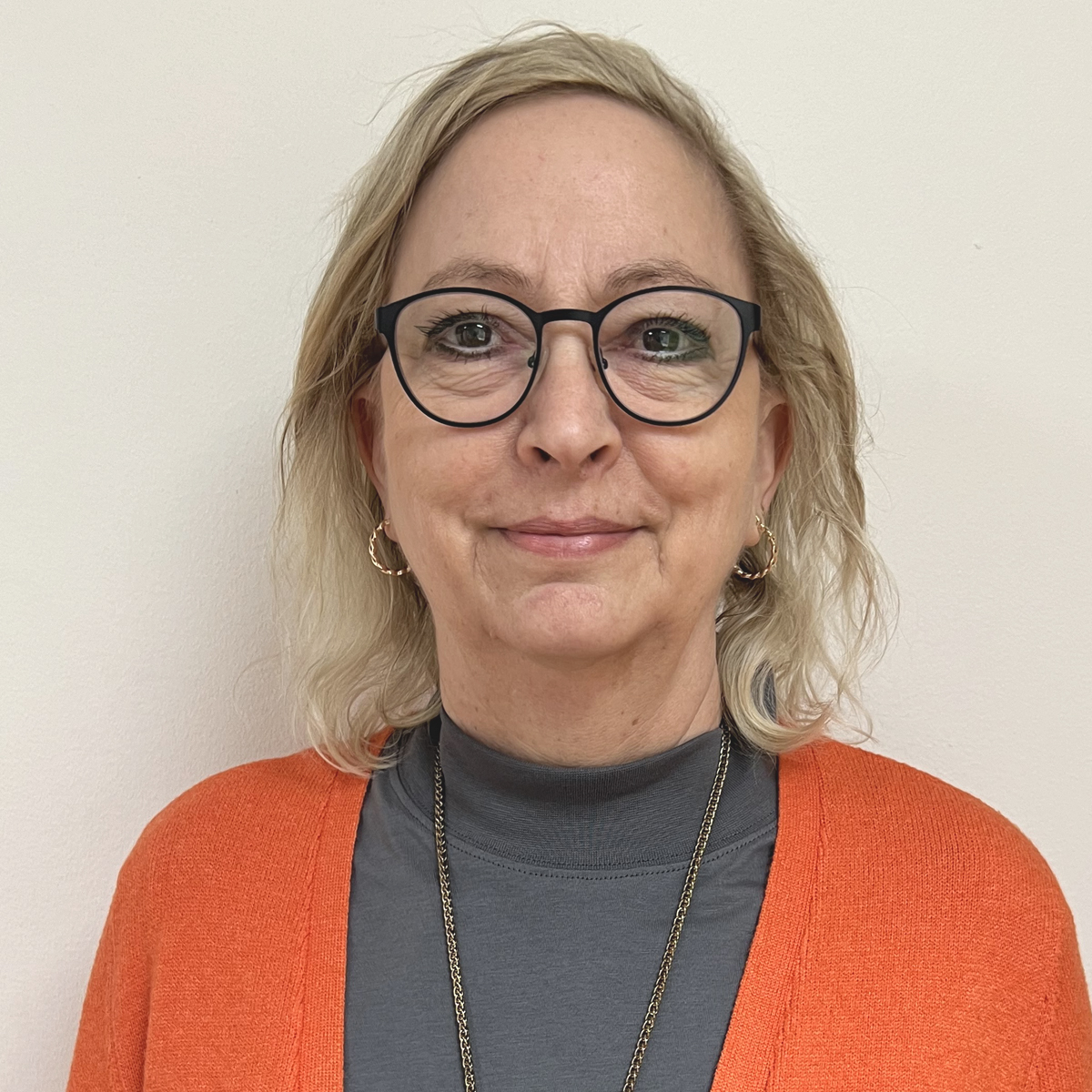 Kvinna i 50-årsåldern, blont axellångt hår, glasögon, orange kofta.