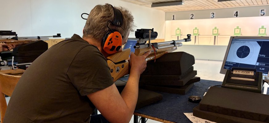 En man siktar med ett luftgevär. Han har hörlurar på sig. Bredvid honom syns en display med måltavlan på. Framför honom finns en vägg med upplysta måltavlor.