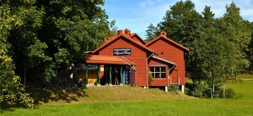 En sommarbild på en röd timmerstuga som är museet. På vänster sida finns stora gråna träd som delvis skymmer stugan