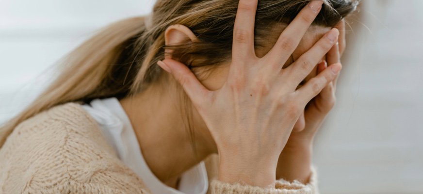 En person sitter framåtlutad och håller händerna mot ansiktet så att ansiktet nästan helt täcks. Håret är uppsatt bakåt i en hästsvans och personen bär en ljus stickad tröja. Bilden är en närbild och ger ett intryck av stress eller oro.