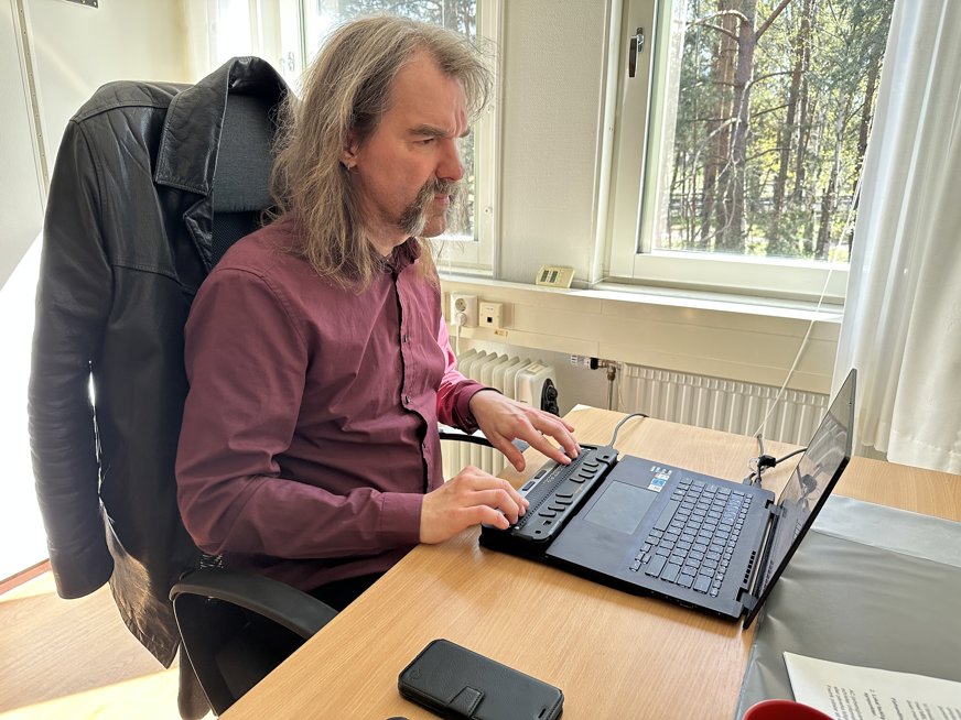Fredrik sitter vid ett skrivbord och skriver på en punktdisplay framför en bärbar dator. I bakgrunden syns ett fönster med träd utanför.