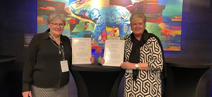 Två leende kvinnor i 50-årsåldern vid ståbord med varsitt diplom, i bakgrunden stor kulört tavla.