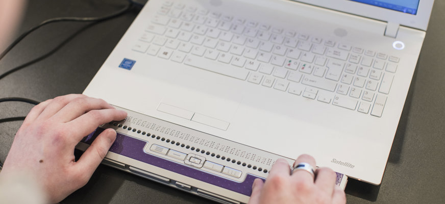 Händer som skriver på en punktskriftsdisplay kopplad till en vit dator, som står på ett svart bord.