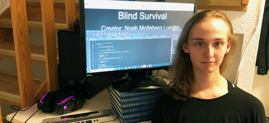 Noah har långt hår, en svart tröja och tittar in i kameran. I bakgrunden syns en skrivbordsplats med datorskärm, ett par hörlurar och högtalare. Den är placerad under en trappa.