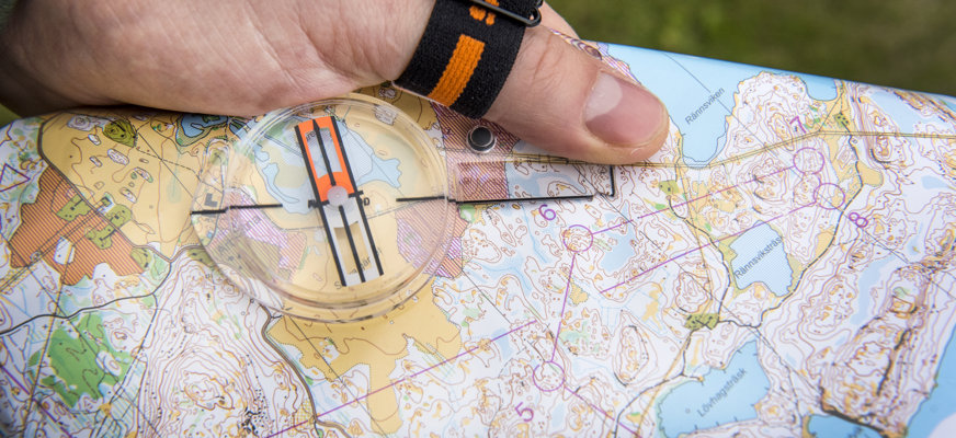 En bild på en terrängkarta och en hand som håller i den. På kartan ligger en kompass. 
