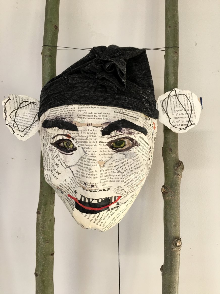 Denna mask är tillverkad av tidningspapper, vilket skapar en collage-effekt på ytan med synliga textstycken som bidrar till dess unika utseende. Masken har ett uttrycksfullt ansikte med markerade, mörka ögonbryn som ger den en dramatisk framtoning. Ögonen har en intensiv blick, och irisar målade i en gröngul ton framträder tydligt mot det svartvita pappret. Maskens mun är ett enkelt, rött leende, vilket ger ansiktet ett vänligt eller nöjt uttryck. Masken bär en mörk huvudbonad av tyg, som är draperad på ett sätt som lägger till en extra dimension och textur. Öronen, gjorda av papper och dekorerade med handritade linjer, står ut från maskens sidor och bidrar till den humoristiska, lite lekfulla känslan. Masken är monterad mellan två bambustänger, vilket ger ett naturligt intryck, och kombinationen av tidningstext, färg och tyg ger verket en känsla av personlighet och historia.