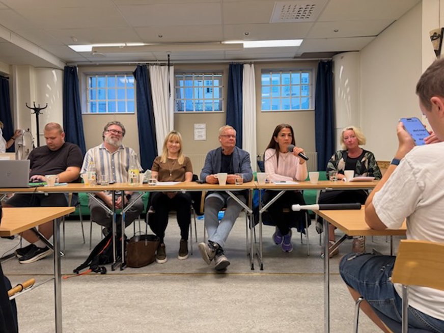 Från debatten 25 aug. 6 personer sitter vid ett bord (podium) längst fram i ett rum, bakom dem 3 fönster med gardiner. Personerna vid bordet är Malin Frick från Centerpartiet, Robert Mjörnberg från Vänsterpartiet, Andréa Hedin från Moderaterna, Gulan Avci från Liberalerna samt Kevin Kjelldahl och Urban Fernquist från SRF Stockholms Stad.