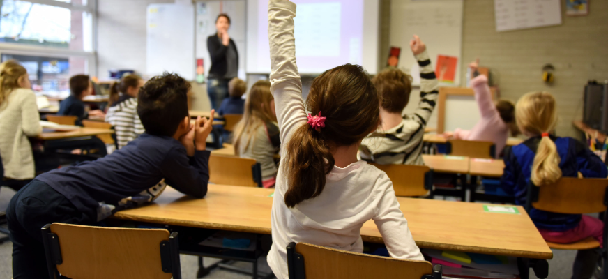 En skolklass sitter vid bord i ett klassrum. Längst fram står en lärare framför en projektorduk. Flera elever räcker upp handen. I förgrunden sitter en flicka med brunt hår uppsatt i en hästsvans och räcker upp ena handen.