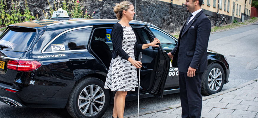 En kvinna och en man står bredvid en taxibil. Kvinnan har sin vita käpp i handen. Mannen är klädd i taxiuniform.