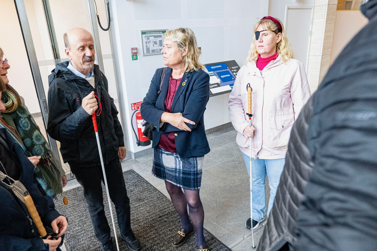 Henrik Eldh håller sin vita käpp till vänster i bild. Birthe Sandberg står i mitten av bilden med armarna i kors över bröstet. Hon har en mörkblå kavaj, rutig kjol och en handväska över högeraxeln. Bakom henne står Helene Jangenfält och lyssnar på samtalet. I bakgrunden syns en taktil karta över Rådhus Skånes lokaler.