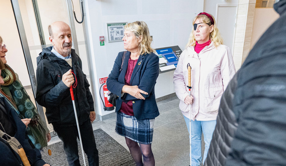 Henrik Eldh håller sin vita käpp till vänster i bild. Birthe Sandberg står i mitten av bilden med armarna i kors över bröstet. Hon har en mörkblå kavaj, rutig kjol och en handväska över högeraxeln. Bakom henne står Helene Jangenfält och lyssnar på samtalet. I bakgrunden syns en taktil karta över Rådhus Skånes lokaler.