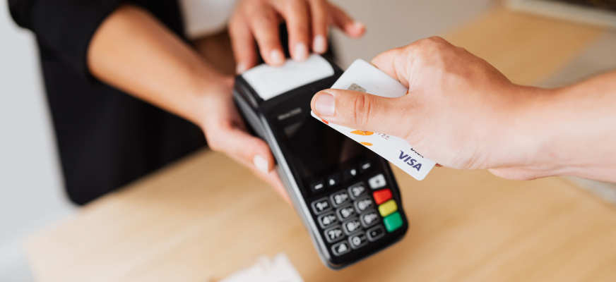 En person håller fram ett bankkort mot en kortterminal. En annan person håller i kortterminalen. Terminalen står på ett träbord och har en knappsats med siffror och färgade knappar. På kortet syns texten ”VISA”.