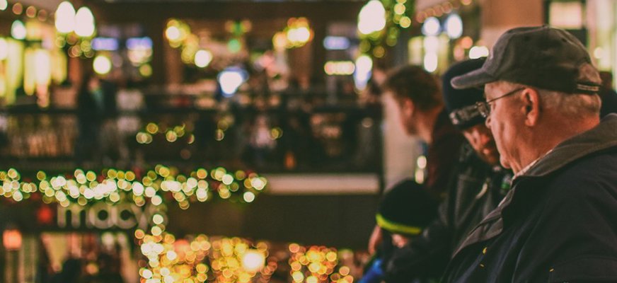 En rymlig hall, det kan vara ett köpcentrum, är julpyntad med ljusslingor och en stor krans i fönstret. I förgrunden syns en man med keps och glasögon.