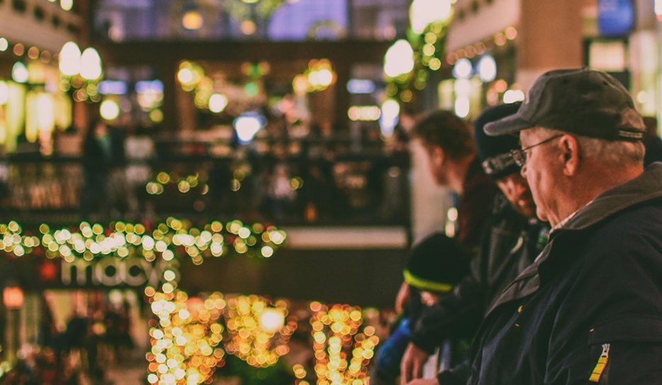 En rymlig hall, det kan vara ett köpcentrum, är julpyntad med ljusslingor och en stor krans i fönstret. I förgrunden syns en man med keps och glasögon.