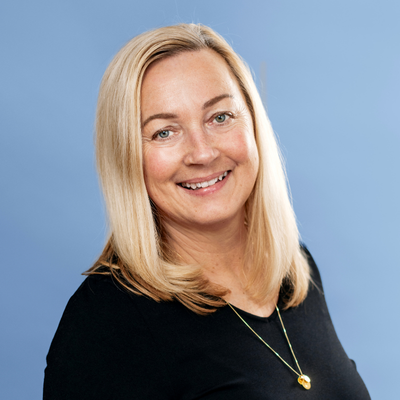 En leende kvinna med axellångt blont hår, iklädd en svart långärmad tröja och ett guldfärgat halsband hänger runt hennes hals. Med lätt böjt huvud tittar hon rakt in i kameran. Bakgrunden går i en ljusblå ton.