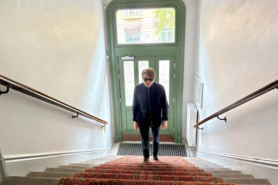 En slank 51-årig vit man med kort brunt hår och mörka glasögon, går upp för en trappa i en äldre trappuppgång. Porten är grön, med utsikt mot gata. Han har svart jacka och byxa