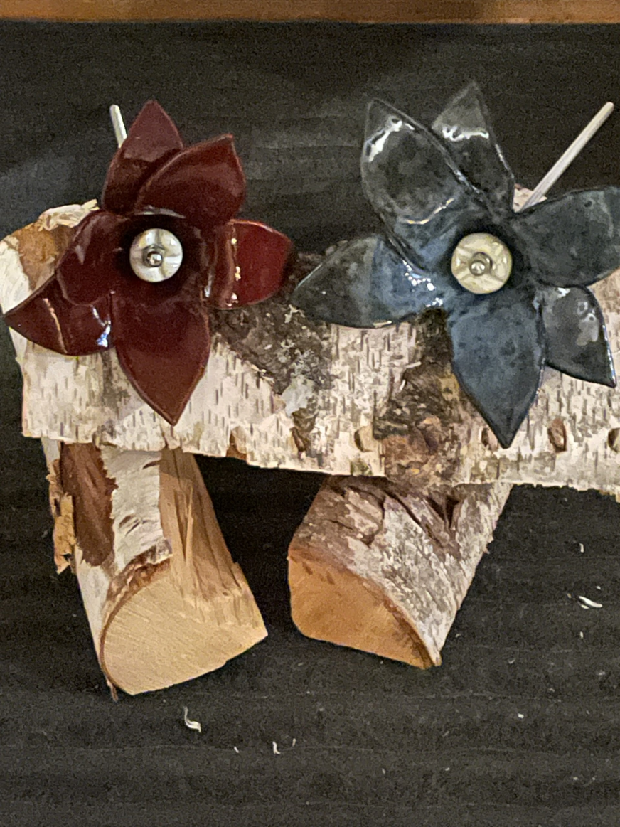 Konstverket består av två blomskulpturer monterade på ett stycke björkträ med delvis bevarad bark. Blommorna har sex glansiga kronblad vardera – en i djupt rött och den andra i mörkt blågrått – och i mitten finns en metallisk detalj som liknar en skruv, vilket ger ett industriellt uttryck. De är fästa med metallstänger direkt i träet, vars naturliga struktur och borrade hål skapar en rustik kontrast till de blanka blommorna. Kombinationen av naturmaterial och mekaniska inslag ger verket en unik balans mellan organiskt och konstruerat, där enkelhet möter precision.