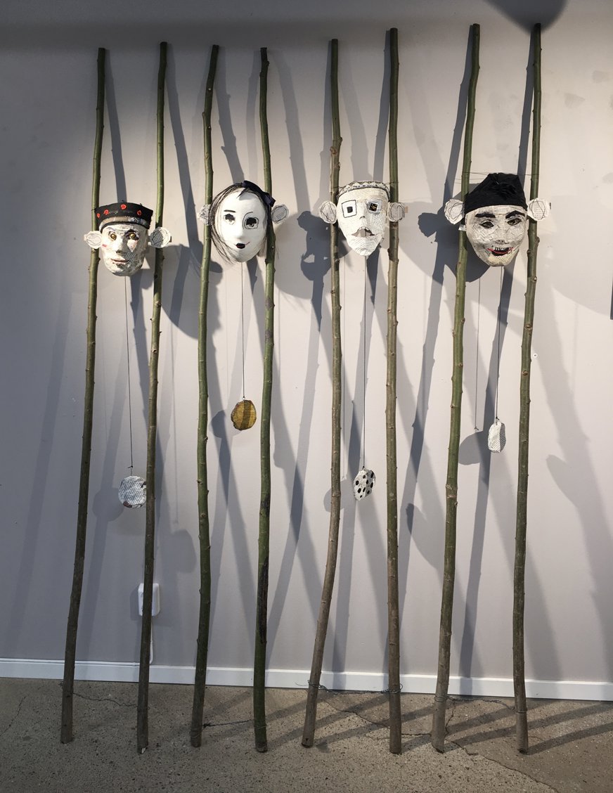 Det här konstverket består av fem masker som är upphängda på långa, tunna stavar av bambu eller liknande naturligt material. Varje mask har en unik ansiktsdesign och uttryck, och visar en variation av stilar och känslor. Från vänster till höger: Den första masken har ett ansikte målat i jordnära toner med runda, vidöppna ögon och ett svart pannband dekorerat med röda cirklar. Den andra masken är målad i vitt, med mörka ögon och ett förvånat uttryck, som om den blivit tagen på sängen. Den tredje masken har ett rektangulärt ansikte med mustasch och ett ögonlapp eller monokel på ena sidan, och ett litet, allvarligt uttryck. Den fjärde masken har en mer abstrakt design med asymmetriska ögon och detaljerad skuggning, vilket ger den ett lite udda utseende. Den femte masken har ett leende uttryck, med överdrivet röda läppar och en mörk mössa, vilket ger den ett glatt intryck. Nedanför varje mask hänger små, texturerade föremål från snören fästa på stavarna, vilket ger en extra dimension till verket. Skuggorna som maskerna och stavarna kastar på väggen skapar ett djup i bilden. Konstverket har en jordnära, organisk känsla och kan antyda teman som individualitet, kulturell identitet eller en representation av olika personligheter.