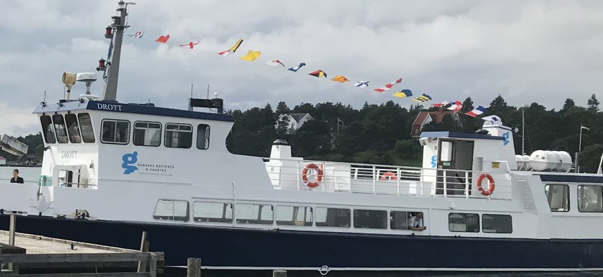 Bild på båten Drott med vit överbyggnad och mörkblå skrovkant. Namnet syns framme vid bryggan och uppe på styrhytten. På sidan finns texten “Gunnars Båtturer & Charter” och en blå logotyp. Längs taket löper en rad färgglada flaggor i rött, gult, blått och vitt. På däck finns flera orange livbojar. I nederkanten av skrovet sitter en rad stora fönster längs hela sidan. I bakre delen syns ytterligare en överbyggnad med dörr och fönster. I bakgrunden syns villor bland trädklädda höjder på Stenungsön.