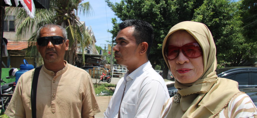 Ramli i kort grått hår, solglasögon och beige skorta, Nur i kortklippt mörkt hår och vit skjorta och vänder sidan mot kameran. Eriwati har beige huvudduk och rödtonade glasögon. Hon är klädd i en brun- och vitrandig topp. Bakom dem syns gatumiljön.