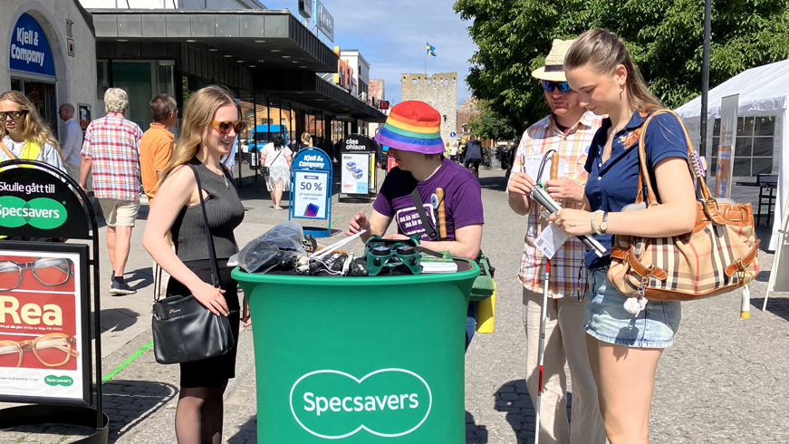 En ung kvinna visar fingerad optik för några yngre vid ett grönt Specsavers-podium.