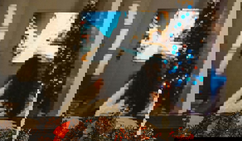 En person sett rakt bakifrån som sitter framför en tv. Bredvid tv:n står en pyntad julgran och i förgrunden syns ett tänt ljus, röda band och kottar.