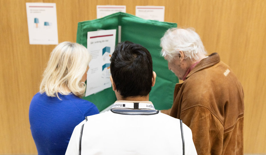 Äldre man i röstningsbås. Två personer assisterar honom.