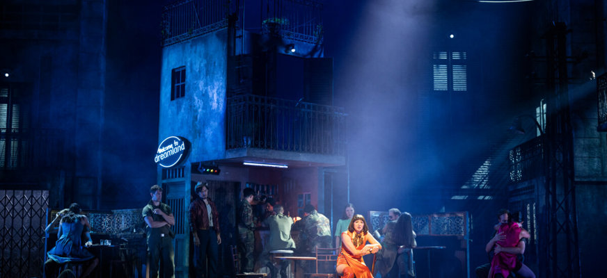 En scen ur musikalen, en kvinna är i strålkastarljuset, resten av scenen är väldigt mörk, man skymtar byggnader bakom henne.